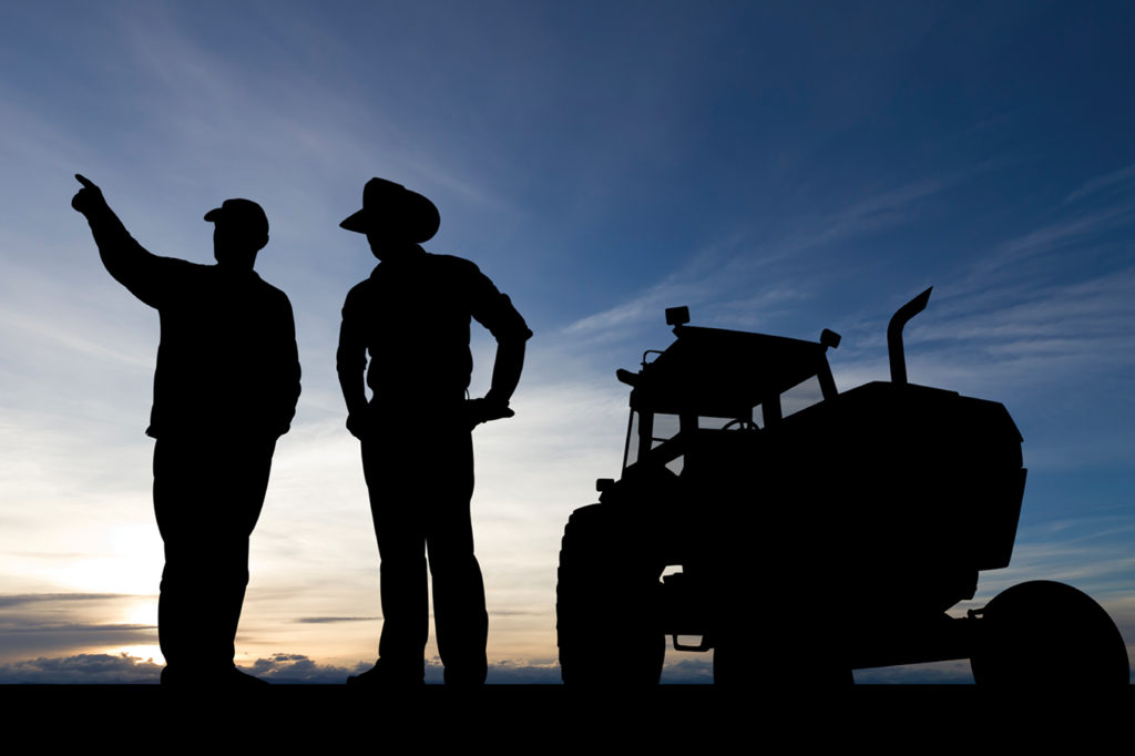 Silhouettes of two farmers looking over fields with tractor behind them